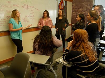 A group of students standing in a circle talking in front of a whiteboard 