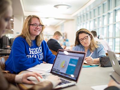 A group of students working at a table on computers and smiling 